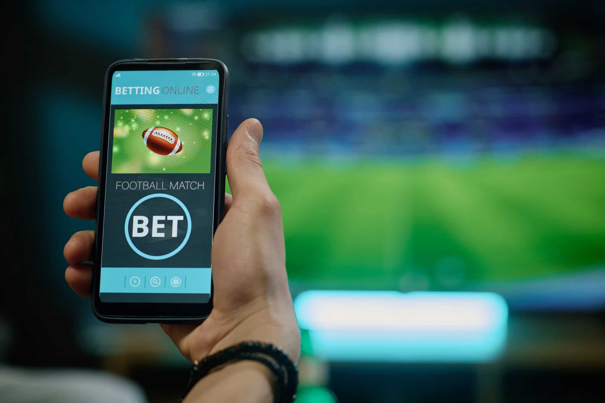 Ao fundo, borrado, uma partida de futebol passando em uma TV; à frente, a foto de uma bola de futebol americano e a palavra "bet" em um smartphone