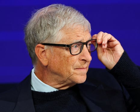 Bill Gates adjusts his glasses, wearing a dark sweater against a blue background