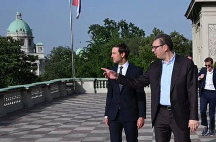 Kushner and Vučić stand on an outdoor roof terrace; Vučić appears to be pointing at something. There is a tall building with a green dome in the background and a Serbian flag at the balustrade.