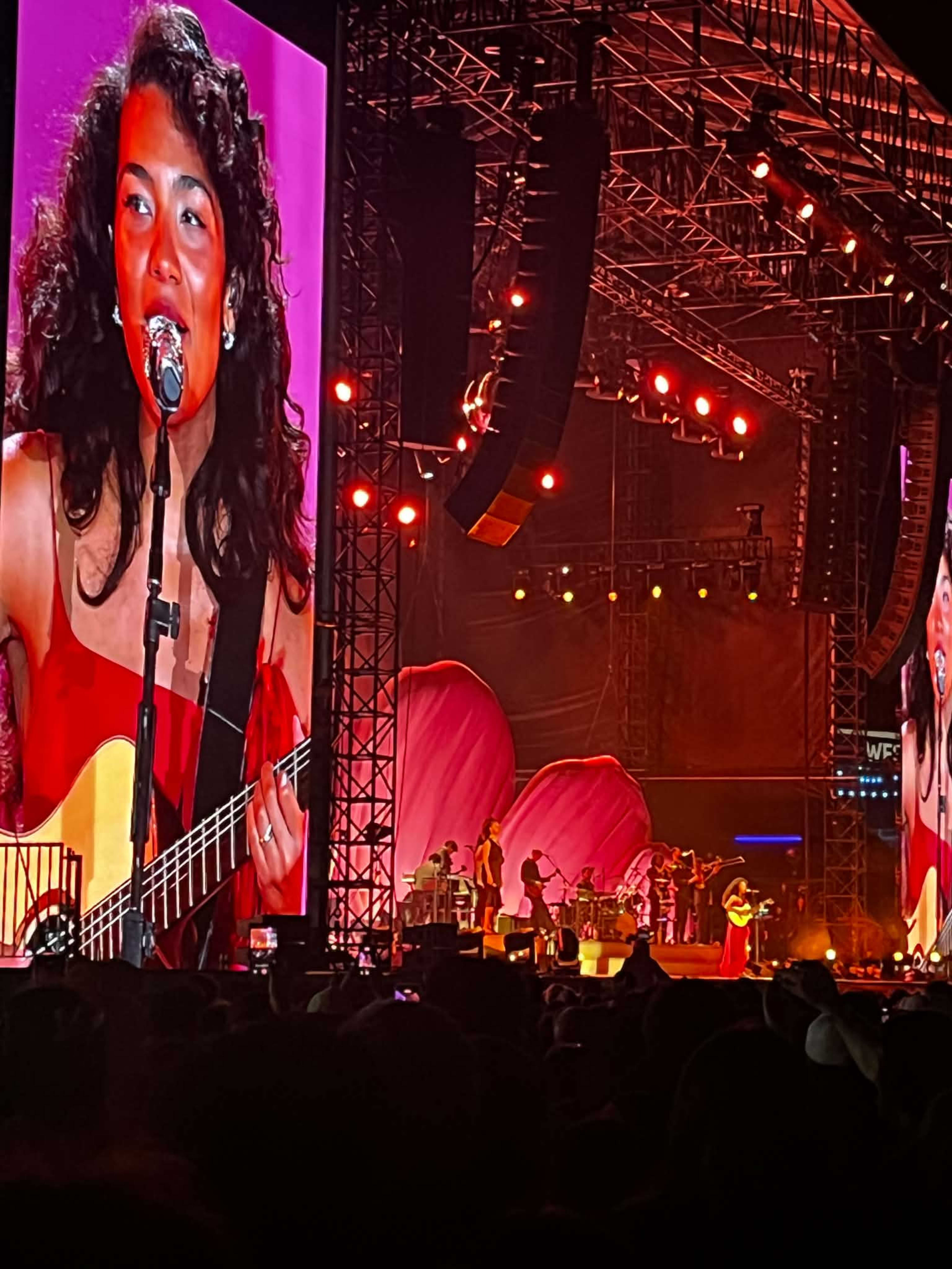 Olivia Dean appears on the big stage screen at Western Springs Bowl as she performs in a red dress while playing the guitar and singing.