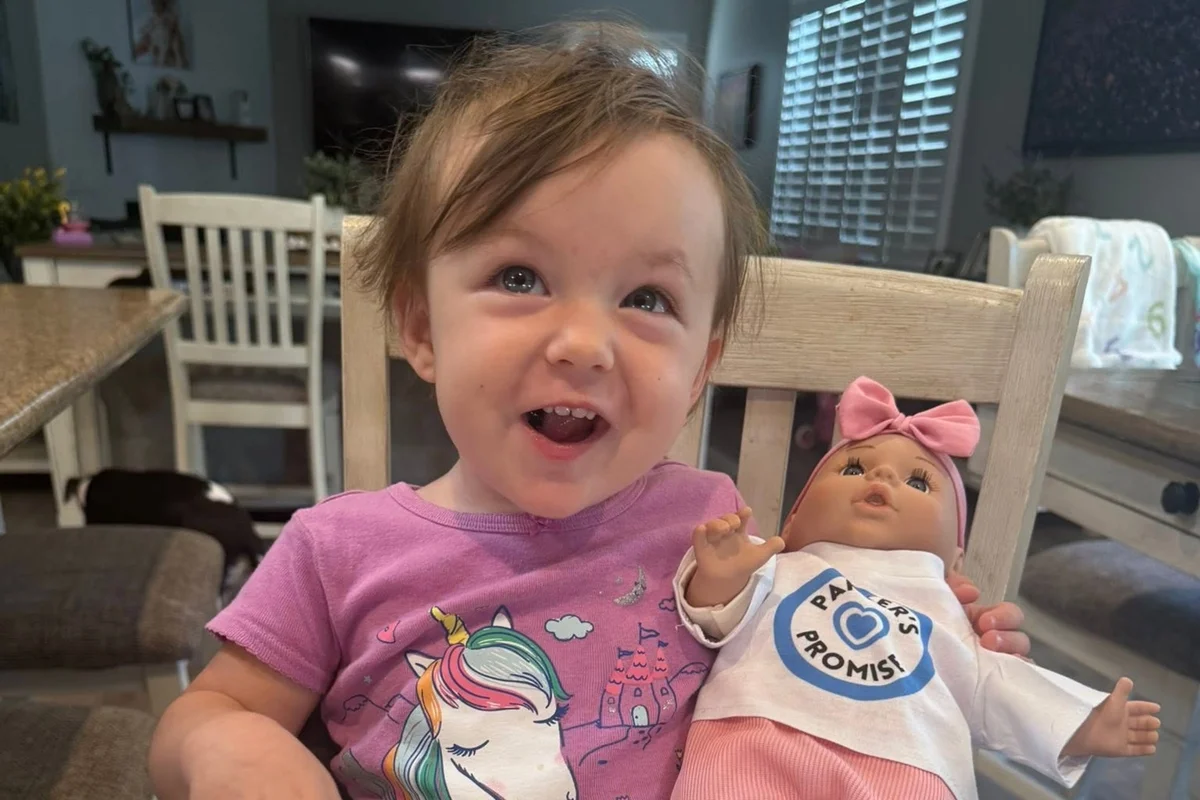 Foto colorida de bebê sorrindo para foto enquanto segura uma boneca - Metrópoles.