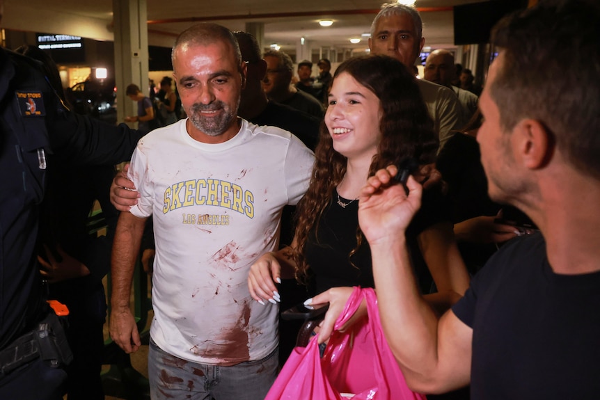 A man, with blood on his tshirt, walks through an airport terminal with a security officer and woman by his side.